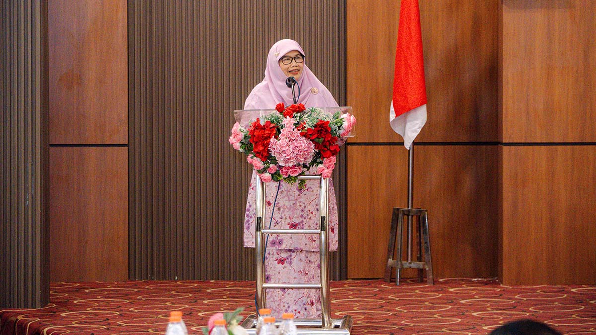 Pembicara berhijab ungu muda menyampaikan sambutan di podium berhias bunga, di samping bendera Merah Putih.