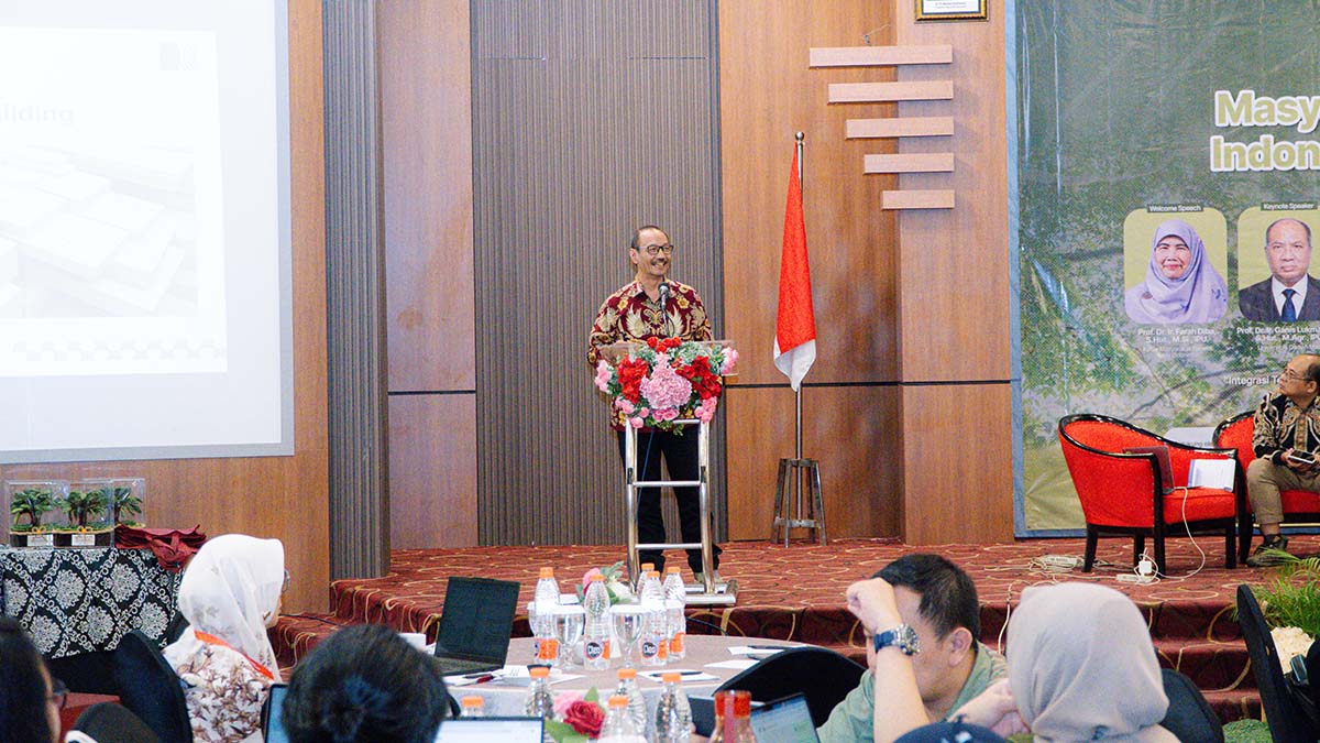 Pembicara berbaju batik menyampaikan materi di podium, dengan layar presentasi dan peserta di ruang seminar.
