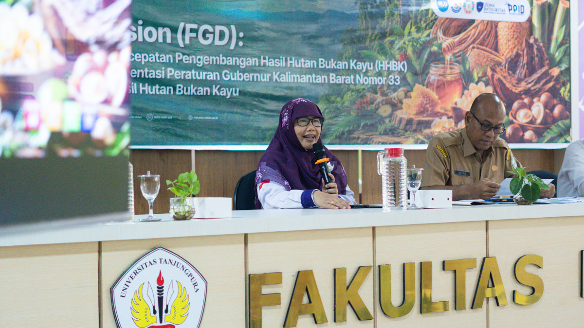Seorang pembicara berjilbab ungu menggunakan mikrofon di meja depan dengan latar spanduk FGD Pokja HHBK.