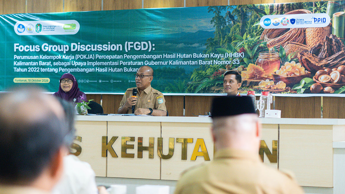 Bapak Adi Yani berseragam dinas cokelat menyampaikan keynote menggunakan mikrofon di meja depan dengan latar spanduk FGD Pokja HHBK.