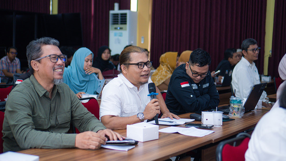 Seorang peserta berkemeja putih berbicara menggunakan mikrofon sambil duduk bersama peserta lain di meja diskusi.