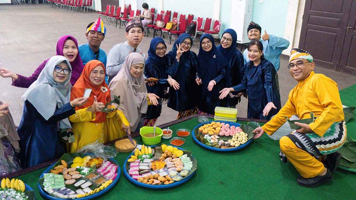 Peserta berbaju Melayu berkumpul mengelilingi hidangan kue di atas nampan; beberapa peserta memegang piring dan makanan.